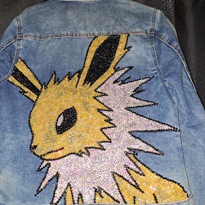 May include: A light blue denim jacket featuring a large, sparkling cartoon character design on the back. The character is yellow and black, with iridescent sequins and black beads creating a shimmering effect. The jacket is hanging on a black hanger.