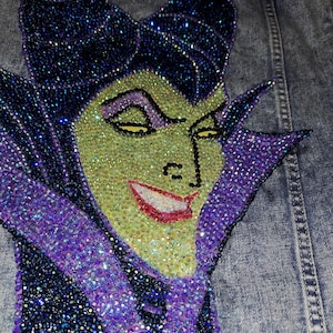May include: Denim jacket with a sparkling, detailed depiction of a character with green skin, purple accents, and black horns. The artwork is made of many small, glittering embellishments.