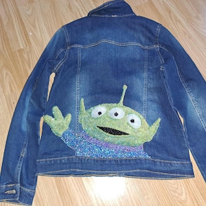Puede incluir: Chaqueta vaquera azul con un diseño de alienígena de tres ojos en la espalda. El alienígena es verde con un cuerpo azul y morado con cuentas. La chaqueta tiene cuello y botones.