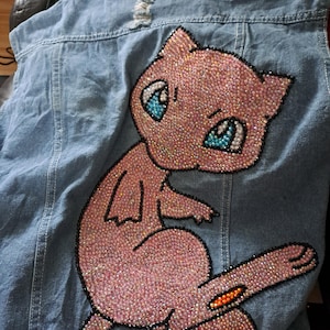 Peut inclure: Veste en jean bleu clair avec un grand personnage de dessin animé rose recouvert de strass au dos. Le personnage a des yeux bleus et est souligné en noir. La veste présente des détails usés.