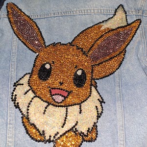 May include: Light blue denim jacket featuring a large, sparkling Eevee design on the back. The Eevee character is embellished with gold, brown, and black rhinestones, creating a shimmering effect. The jacket has a classic denim style.