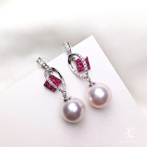 Pode incluir: Um par de brincos de prata com uma pérola branca e uma pedra quadrada rosa. Os brincos apresentam um design delicado e entrelaçado.