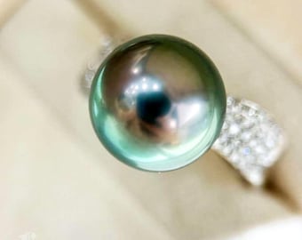 Pode incluir: Um anel com uma pérola grande, redonda e verde escura com uma banda prateada incrustada com pequenos diamantes brancos.