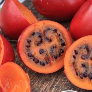 Op de afbeelding: Close-up van tamarillo-vruchten, sommige heel en andere doorgesneden. De schil is felrood, het vruchtvlees oranje en de zaden donker. De vruchten liggen op een houten oppervlak, wat hun unieke vorm en kleur benadrukt.