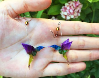 May include: A pair of iridescent origami hummingbird earrings with gold accents. The earrings are made of paper and are suspended from silver hooks.