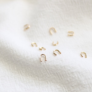 May include: A collection of small, gold, U-shaped hoops, scattered on a white fabric surface.