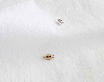 May include: Two small magnetic clasps on a white background. One clasp is gold-colored, and the other is silver-colored. The clasps are cylindrical with small loops on each end. The background is a textured white fabric.