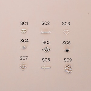 May include: Nine different silver charm pendants, labeled SC1 through SC9. The charms include a heart, a cross, a flower, a circle with a rhinestone, a circle with a black stone, a double loop, a bar, and two different shapes with loops.