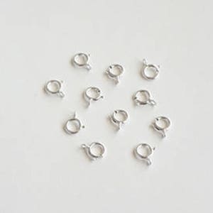 May include: A group of silver-toned metal jump rings, also known as split rings, on a white surface. These rings are used for connecting jewelry components.