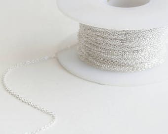 May include: A spool of silver chain, with a portion of the chain unraveled and lying on a white surface.