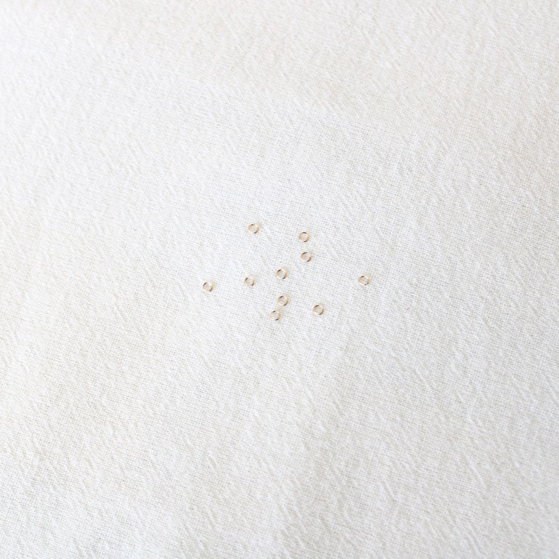 May include: A collection of small, gold jump rings scattered on a white fabric surface.