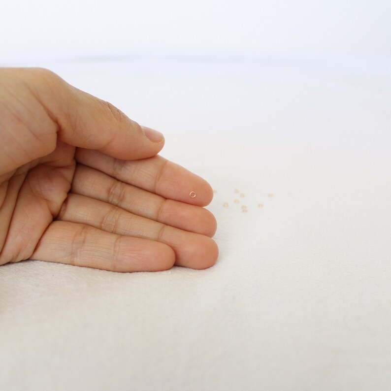 May include: A small gold jump ring, about the size of a grain of rice, is held in a hand. The ring is a common jewelry finding used for connecting chains, charms, and other jewelry components.