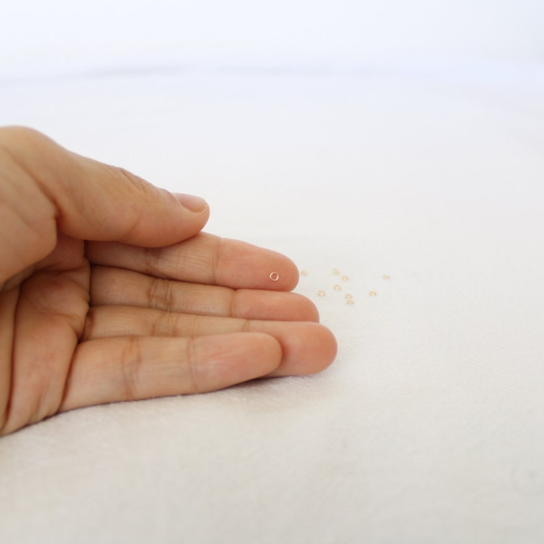 May include: A small gold jump ring, about 4mm in diameter, is held in a hand. A scattering of tiny gold beads are on a white surface.