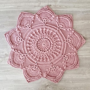 May include: A handmade, dusky pink crochet rug in a floral design. The rug features a central circular pattern surrounded by petal-shaped edges. The intricate crochet work includes textured details and raised elements, creating a decorative and tactile piece.
