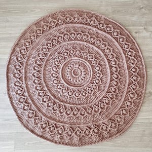 May include: A round, dusty rose crochet rug with intricate, repeating patterns. The rug features concentric circles of textured designs, including floral and geometric motifs. The handmade rug is suitable for a living room or bedroom.
