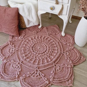 May include: A handmade crochet rug in a dusty rose hue, showcasing a detailed floral pattern. The rug's design includes a central circular motif and petal-shaped borders. The textured surface, created by the crochet, adds a bohemian aesthetic to the space.