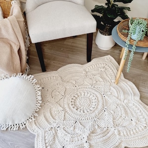 Puede incluir: Una alfombra de ganchillo hecha a mano de color crema, con un borde festoneado y un intrincado diseño floral. La alfombra está colocada sobre un suelo de madera, cerca de una silla blanca y un cojín redondo con flecos.