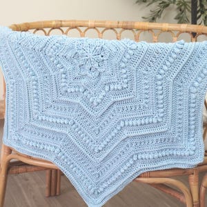 Puede incluir: Una manta de ganchillo azul claro con un diseño de estrella en el centro, colocada sobre una silla de mimbre. La manta presenta intrincados patrones de puntadas y una superficie texturizada. El color azul suave de la manta resalta la artesanía.