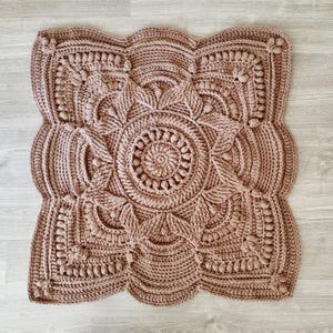 Puede incluir: Un cuadrado de ganchillo hecho a mano y detallado en un color rosa apagado. El cuadrado presenta un diseño circular central rodeado de intrincados patrones florales y geométricos. Los bordes tienen un ribete festoneado.