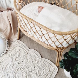 Pode incluir: Um beb&eacute; dorme tranquilamente num ber&ccedil;o de vime tecido, coberto por um cobertor branco. Um tapete de croch&ecirc; creme com um desenho floral est&aacute; no ch&atilde;o de madeira. Uma planta em vaso &eacute; vis&iacute;vel.