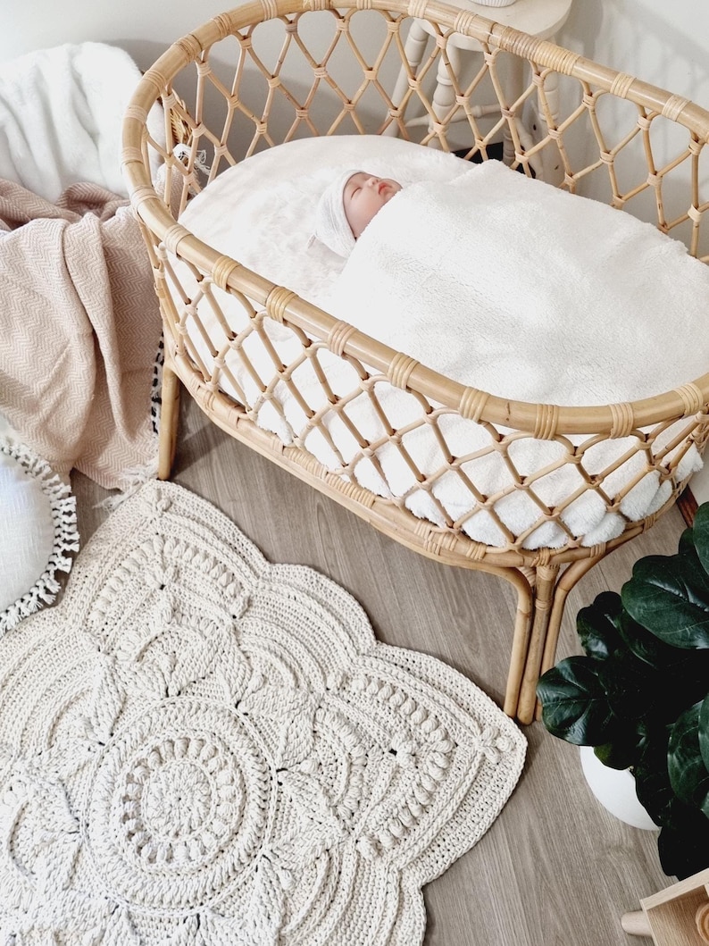 Pode incluir: Um beb&eacute; dorme tranquilamente num ber&ccedil;o de vime tecido, coberto por um cobertor branco. Um tapete de croch&ecirc; creme com um desenho floral est&aacute; no ch&atilde;o de madeira. Uma planta em vaso &eacute; vis&iacute;vel.
