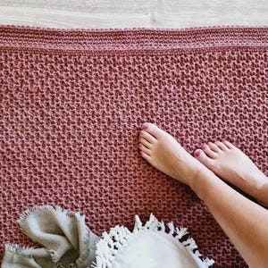 May include: A handmade, dusky pink crochet blanket with a textured stitch pattern. The blanket has a solid border and is shown with a grey scarf and a white, fringed decorative item. The blanket is on a light wood surface.