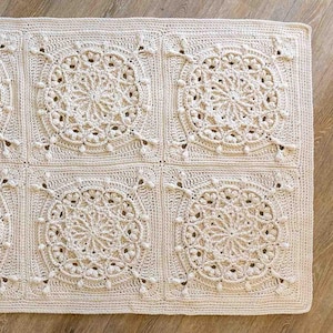 Puede incluir: Manta cuadrada de ganchillo de color crema con un patrón de medallón floral repetido. La manta está hecha de un hilo suave y texturizado y tiene un diseño calado detallado. Un artículo de decoración del hogar acogedor.