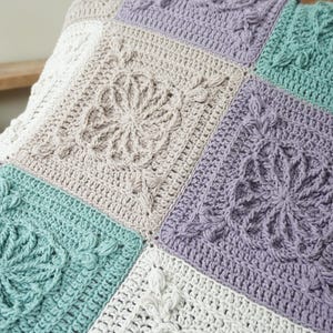 Puede incluir: Una manta de ganchillo hecha a mano con un diseño de patchwork. La manta está compuesta por paneles cuadrados en varios colores pastel, incluyendo lavanda, verde azulado y crema. Cada cuadrado tiene un patrón floral en relieve, agregando textura al textil suave y acogedor.