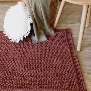 May include: A burgundy hand-knitted rug with a textured pattern and a thick border. A white fringed cushion and a woven basket with a grey blanket are in the background. A small wooden side table is also visible.