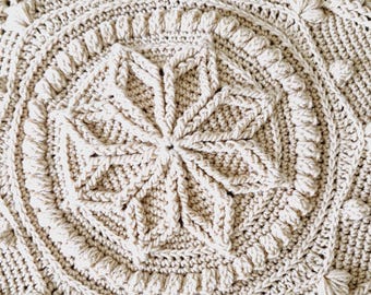 Puede incluir: Primer plano de una manta de ganchillo con un diseño detallado e intrincado en color crema. La manta presenta un patrón circular con un motivo central en forma de estrella y un borde texturizado.
