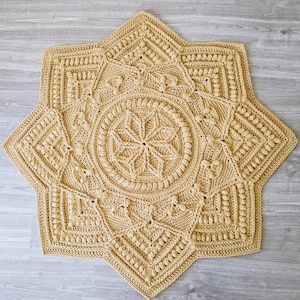 Op de afbeelding: Een beige, stervormig gehaakt kleed met ingewikkelde patronen. Het kleed heeft een centraal bloemmotief omgeven door geometrische vormen en getextureerde steken. Het kleed heeft een diameter van ongeveer 100 cm.