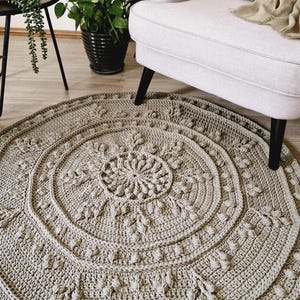 May include: A round, light gray crochet rug with a detailed floral pattern. The rug features concentric circles with textured leaf and bobble stitch designs. The rug is placed on a light wood floor, with a white armchair and plants in the background.
