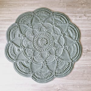 May include: A round, sage green crochet rug with a detailed floral design. The rug features a central circular motif surrounded by intricate patterns and a scalloped edge. The handmade rug is placed on a light wood floor.