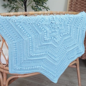 May include: A light blue, handmade crochet blanket with a starburst design. The blanket features a detailed pattern of raised stitches and geometric shapes. It is draped over a wicker crib, showcasing its texture and craftsmanship.