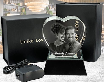 Puede incluir: Un cristal en forma de corazón con una foto grabada en 3D de una pareja. El cristal está iluminado desde abajo con una luz blanca. El texto "Family Forever" está grabado en la base del cristal. El cristal está sentado sobre una base negra.