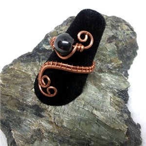 May include: A copper wire ring with a black hematite bead. The ring is wrapped in copper wire and has a swirl design.