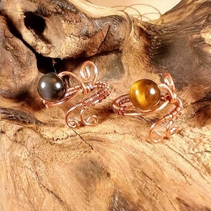 Puede incluir: Dos anillos de alambre de cobre con cuentas de piedras preciosas. El anillo de la izquierda tiene una cuenta negra y el anillo de la derecha tiene una cuenta marr&oacute;n. Los anillos est&aacute;n sobre una superficie de madera.