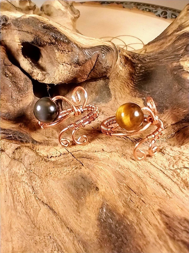 Puede incluir: Dos anillos de alambre de cobre con cuentas de piedras preciosas. El anillo de la izquierda tiene una cuenta negra y el anillo de la derecha tiene una cuenta marr&oacute;n. Los anillos est&aacute;n sobre una superficie de madera.