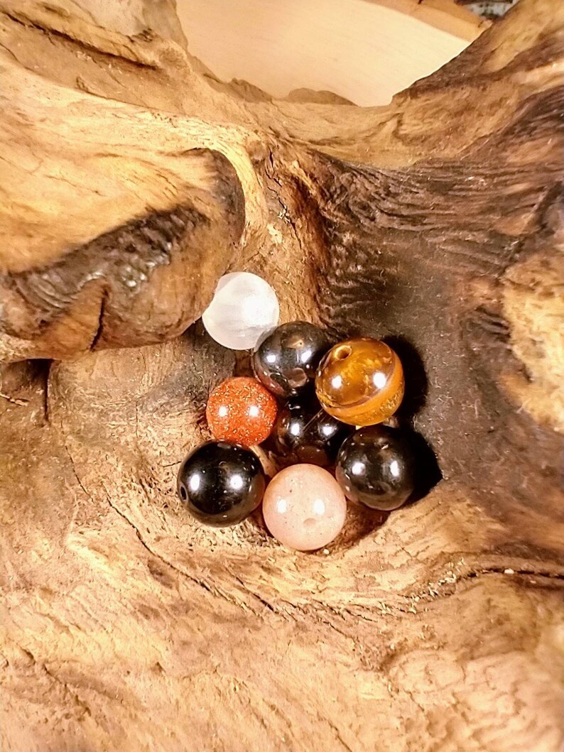 Puede incluir: Una colecci&oacute;n de ocho cuentas redondas de piedras preciosas en varios colores, incluyendo negro, marr&oacute;n, rojo, blanco y beige. Las cuentas est&aacute;n dispuestas en un grupo sobre una pieza de madera.