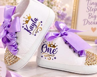May include: A pair of white baby sneakers featuring purple satin ribbon laces, gold rhinestone-encrusted toes, and custom text. One shoe displays the name Kayla with a gold crown, while the other features the word One with a gold crown design.