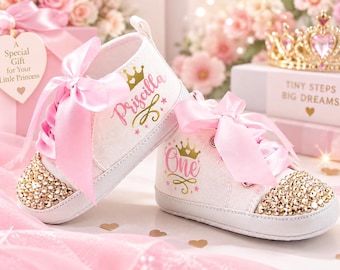 May include: A pair of white baby sneakers with pink satin ribbon laces and gold rhinestone-encrusted toe caps. One shoe features the name Priscilla and a gold crown, while the other displays the word One and a gold crown in pink and gold text.