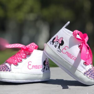 May include: White baby sneakers with pink polka dot laces and a Minnie Mouse graphic. The toe of each shoe is embellished with purple rhinestones. The name "Ember" is printed in pink on the side of each shoe.