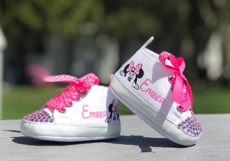 May include: White baby sneakers with pink polka dot laces and a Minnie Mouse graphic. The toe of each shoe is embellished with purple rhinestones. The name "Ember" is printed in pink on the side of each shoe.