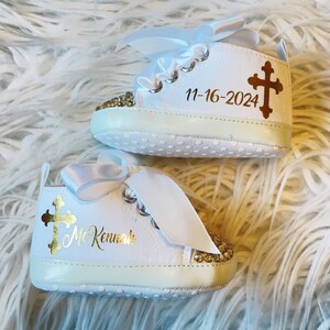 Pode incluir: Sapatos de beb&eacute; brancos com glitter dourado e um design de cruz dourada. Os sapatos s&atilde;o personalizados com o texto "Mr. Kenneth" e a data "11-16-2024".
