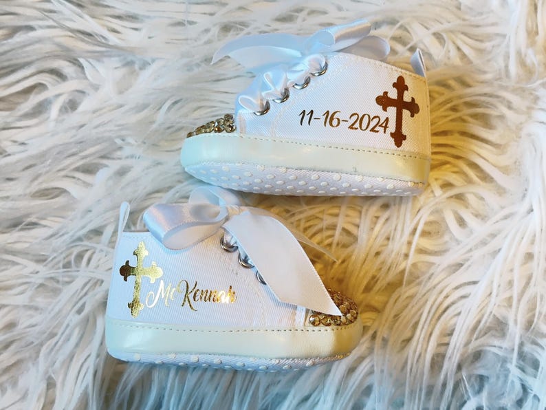 Pode incluir: Sapatos de beb&eacute; brancos com glitter dourado e um design de cruz dourada. Os sapatos s&atilde;o personalizados com o texto "Mr. Kenneth" e a data "11-16-2024".