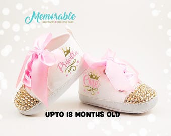 May include: A pair of white baby shoes with pink bows and gold glitter. The shoes are personalized with the names "Priscilla" and "One" and a crown design. The shoes are for babies up to 18 months old.