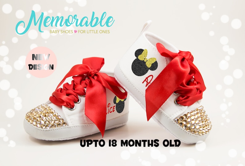 May include: White baby shoes with red satin ribbon laces and a black and gold glitter Minnie Mouse design. The shoes have a gold glitter toe and are embellished with rhinestones. The shoes are for babies up to 18 months old.