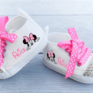 May include: A pair of white baby sneakers with pink polka dot laces and rhinestone-studded toes. Each shoe features a Minnie Mouse graphic and the name "Alice" in pink script. The shoes are designed for infants.