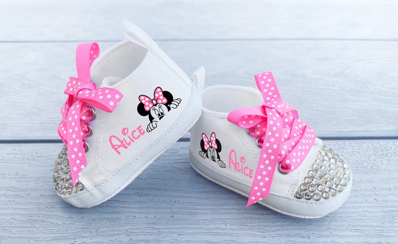 May include: A pair of white baby sneakers with pink polka dot laces and rhinestone-studded toes. Each shoe features a Minnie Mouse graphic and the name "Alice" in pink script. The shoes are designed for infants.