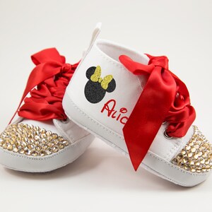 May include: White baby shoes with red satin bows and a black and gold Minnie Mouse design. The shoes are decorated with clear rhinestones and the name "Alice" is written on the side.
